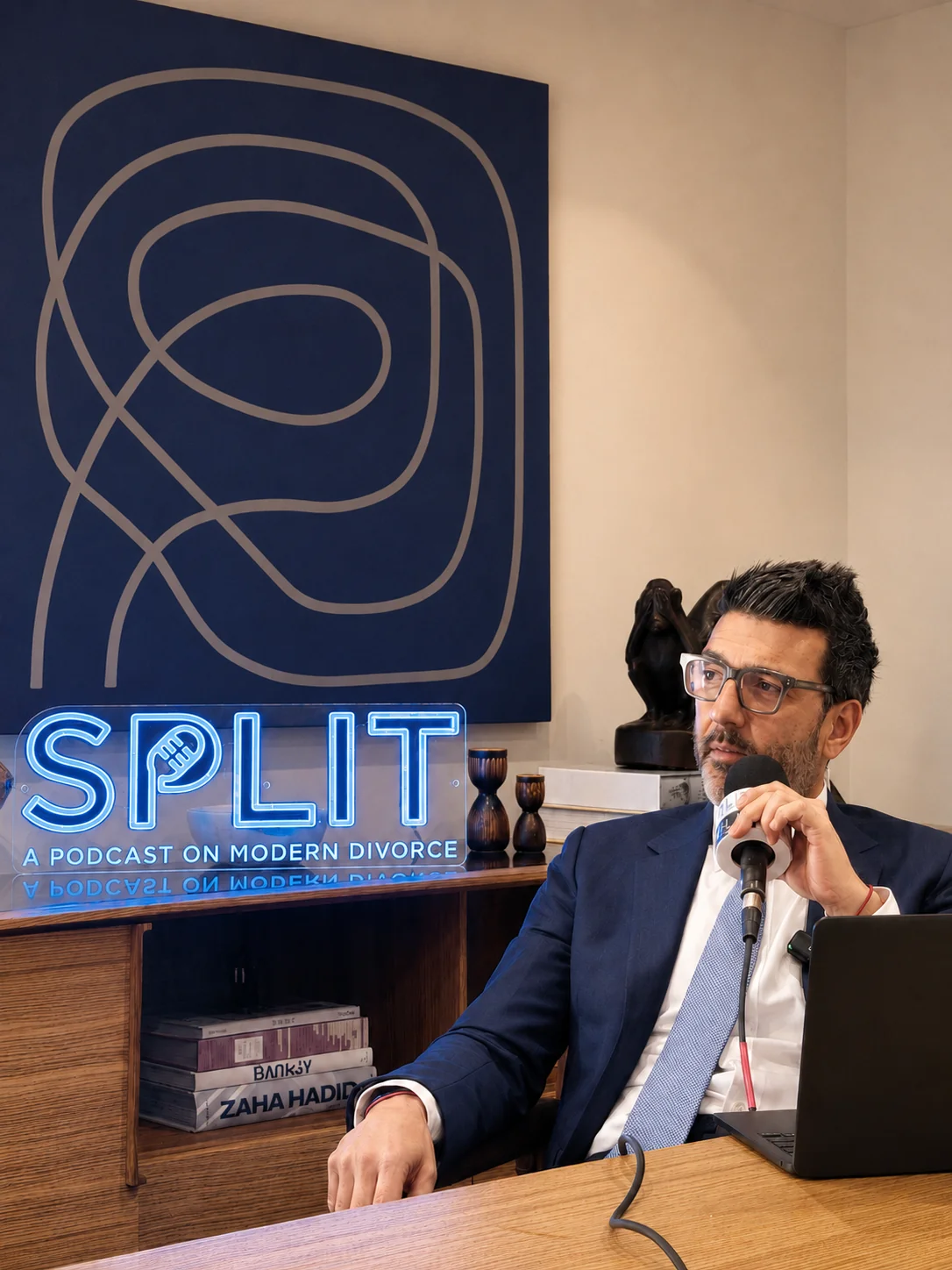 Gus Dimopoulos in the Split podcast studio, behind a microphone, with the neon Split sign and blue artwork in the background.